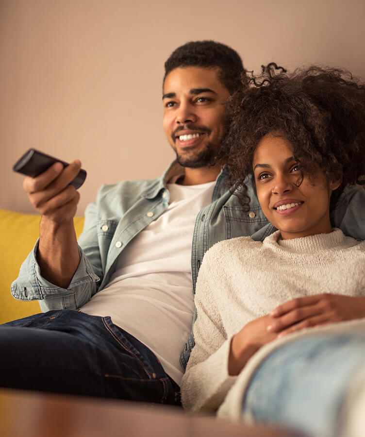 due persone sedute comodamente sul divano e che guardano la TV con una persona che tiene il telecomando
