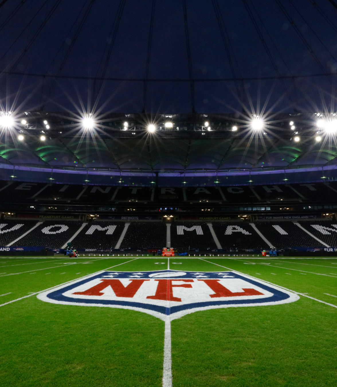 Vista noturna de um campo de futebol americano iluminado da NFL, com o logotipo da NFL no meio do campo dentro de um grande estádio.
